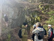 Na fotografii zachycena skupina lidí zastavená v lese u skalnatého útvaru připomínajícího menší jeskyni. P. Neckářová stojí stranou u skály a předčítá zajímavosti o místě, zatímco ostatní účastníci pozorně poslouchají