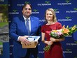 Držitelka ocenění Skutek roku, ředitelka p. Mgr. Bělovová Dolejší, stojí s kyticí květin vedle pana radního Horkýho z Kraje Vysočina. Společně pózují před modrým prezentačním panelem s logem kraje, on drží dárkový balíček a pamětní listinu