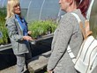 Na fotografii stojí p. Parkosová venku u fóliovníku a poslouchá místní farmářku, která jí ukazuje pěstované květiny a bylinky. Obě ženy si povídají mezi záhony s rostlinami v příjemném slunečném počasí