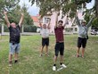 Na fotografii zachyceni zleva: p. Vondra, p. Vlašín, p. Spáčil, p. Svoboda – cvičící na trávníku. Stojí s rukama zvednutýma nad hlavou a provádějí společný protahovací cvik. V pozadí jsou stromy, tráva a obytné domy