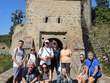 Společná fotografie před historickou kamennou věží hradu Zvíkov. Někteří stojí, jiní sedí na kamenné zídce. Nad nimi je věž s červenou střechou a modrá obloha