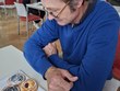 Na fotografii zachycen p. Lukačina během soutěže v Braillově písmu. Sedí u stolu, před sebou má připravené občerstvení a čeká na svůj čas, kdy bude soutěžit. V místnosti jsou další účastníci a stolky s občerstvením