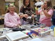 Na fotografii zachyceny dívky, které se zajímají o kompenzační pomůcky TyfloCentra. Kolem procházejí další návštěvníci akce. V pozadí jsou přírodovědné exponáty